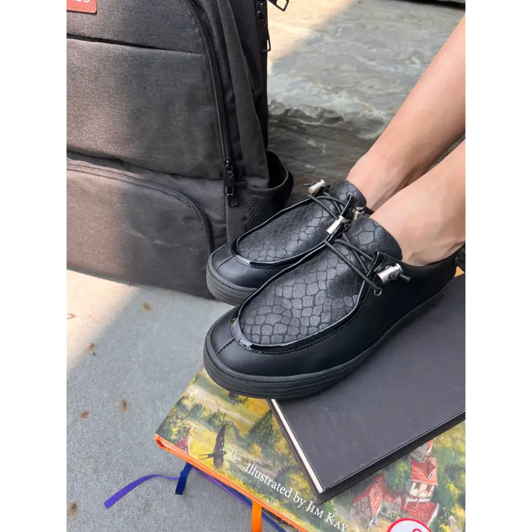 Person wearing black textured shoes with a black bag and book on a concrete surface