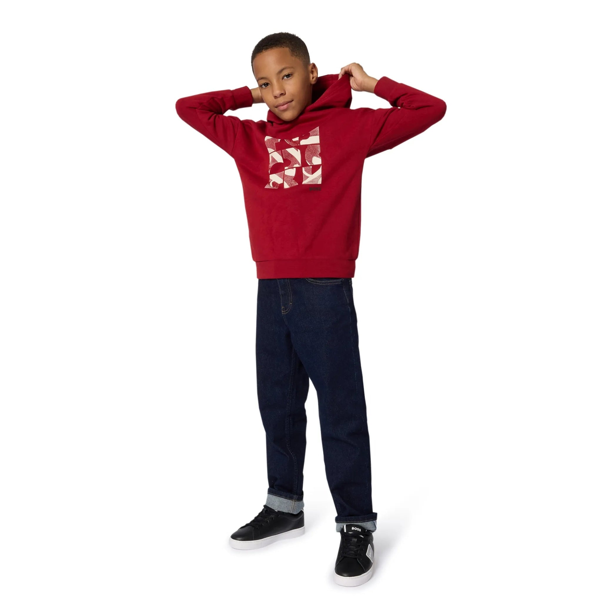 Child wearing a red hoodie with a  pattern, dark jeans, and black shoes on a white background