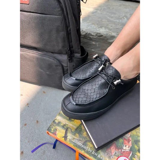 Person wearing black textured shoes with a black bag and book on a concrete surface